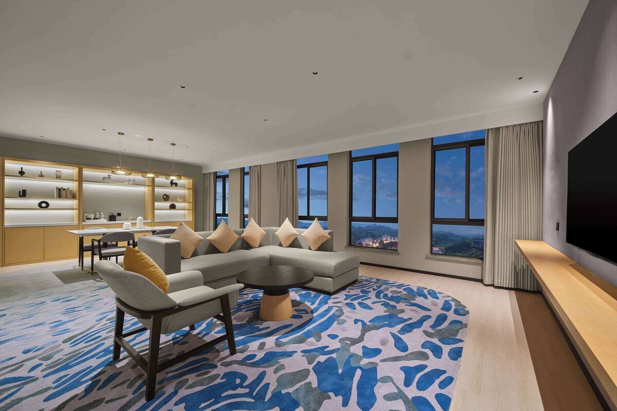 A modern living room with a sectional sofa, a dining area with a table and chairs, a built-in bookshelf, and a large TV screen.