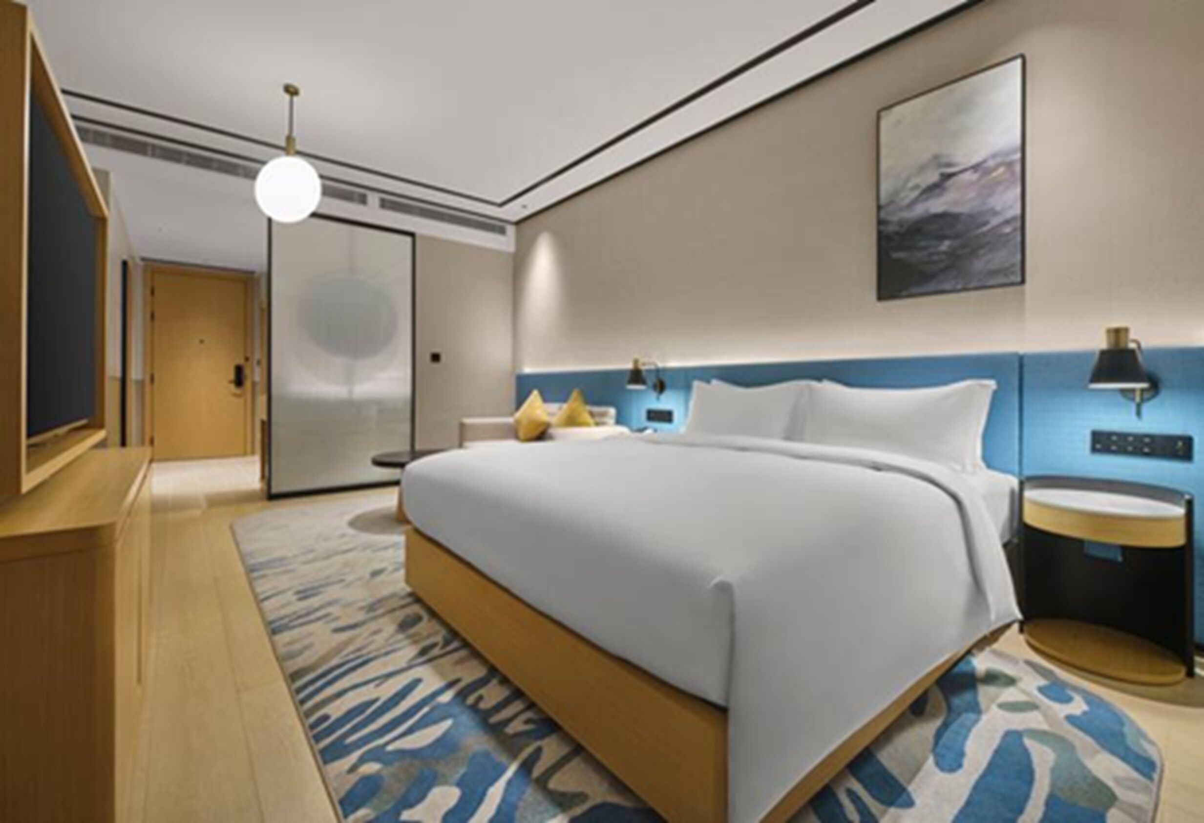 A modern hotel room with a large bed, a desk, a chair, a lamp, and a painting on the wall.