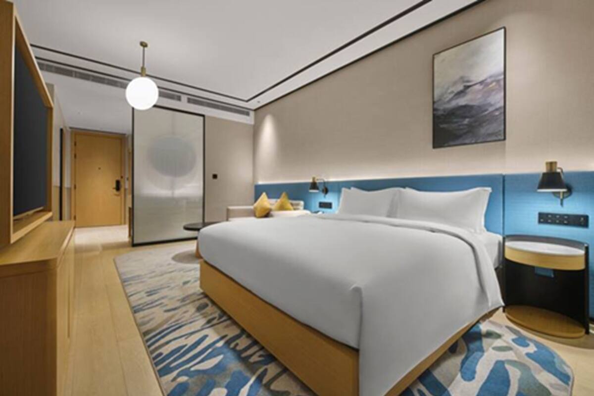 A modern hotel room with a large bed, a desk, a chair, a lamp, and a painting on the wall.