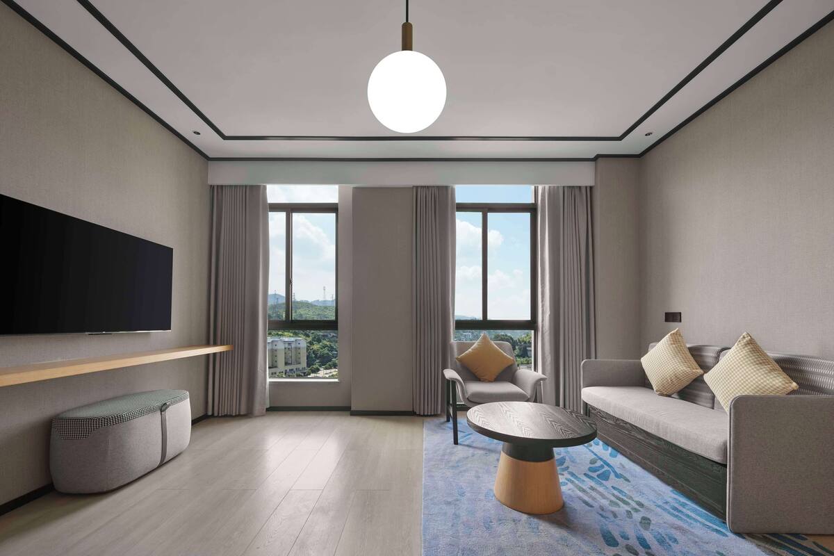 A modern living room with a flat-screen TV, a round coffee table, a sofa with cushions, and a view of a cityscape through large windows.