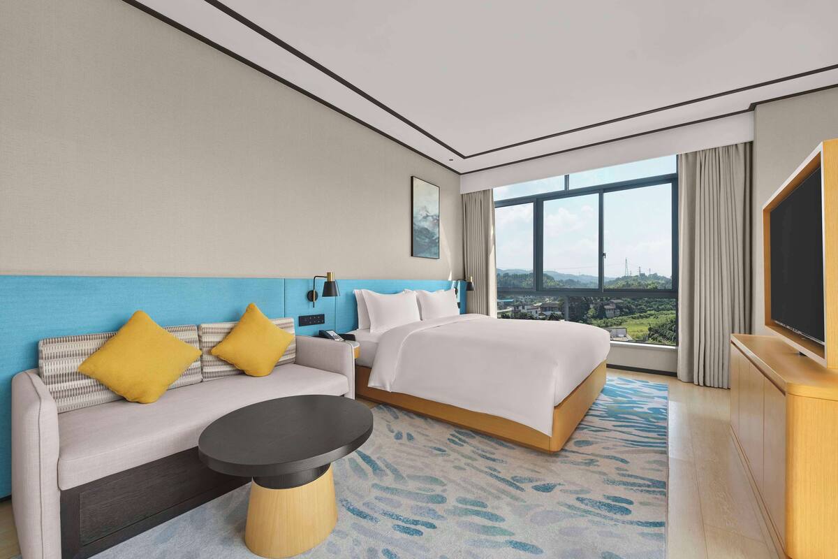 A hotel room with a bed, sofa, and a small table. There is a large window providing a view of the outside.