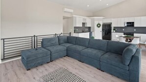 A modern living room with a sectional sofa, a small ottoman, and a dining area with a table and chairs. - Modern 2024 build, in welcoming Lava Hot Springs. Top unit. (Lava Hot Springs)