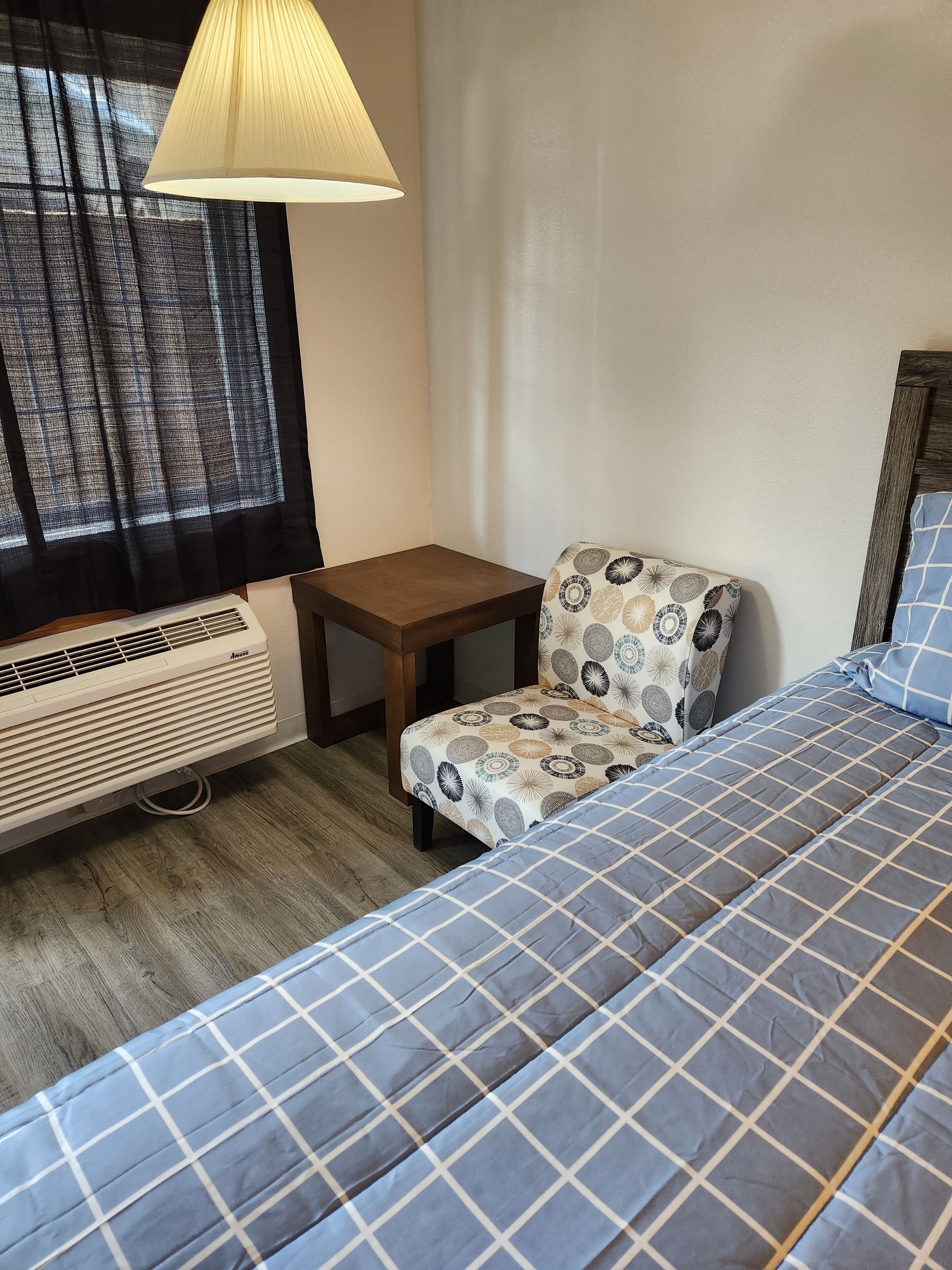 A hotel room with a bed, a chair, a small table, a window with curtains, and an air conditioning unit.