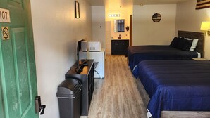 Une chambre d’hôtel avec une porte verte, un bureau avec un ordinateur, une poubelle, un lit avec une literie sombre, une petite kitchenette et un miroir au-dessus du lavabo.