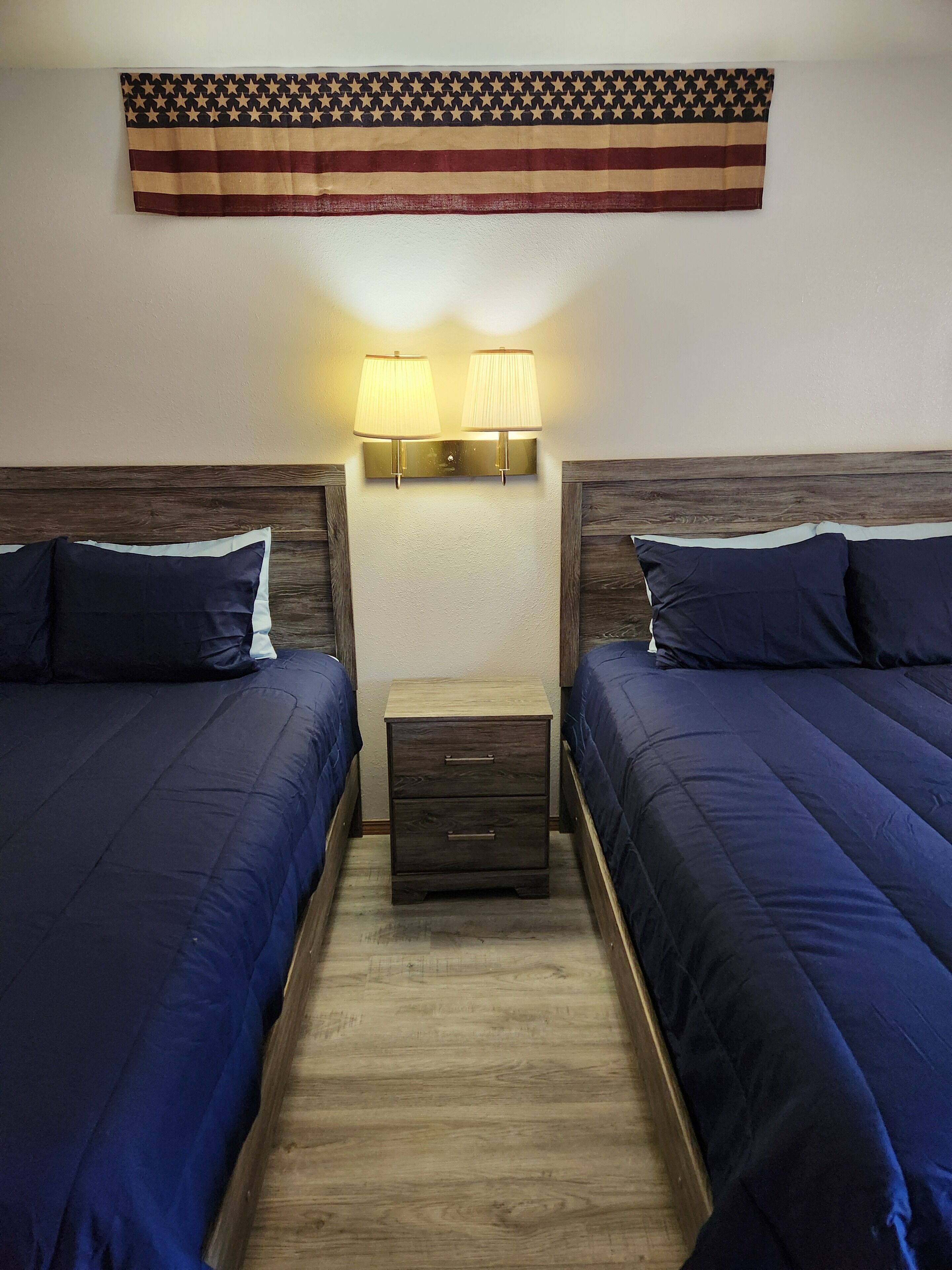 A hotel room with two beds, a nightstand, and a wall-mounted lamp.