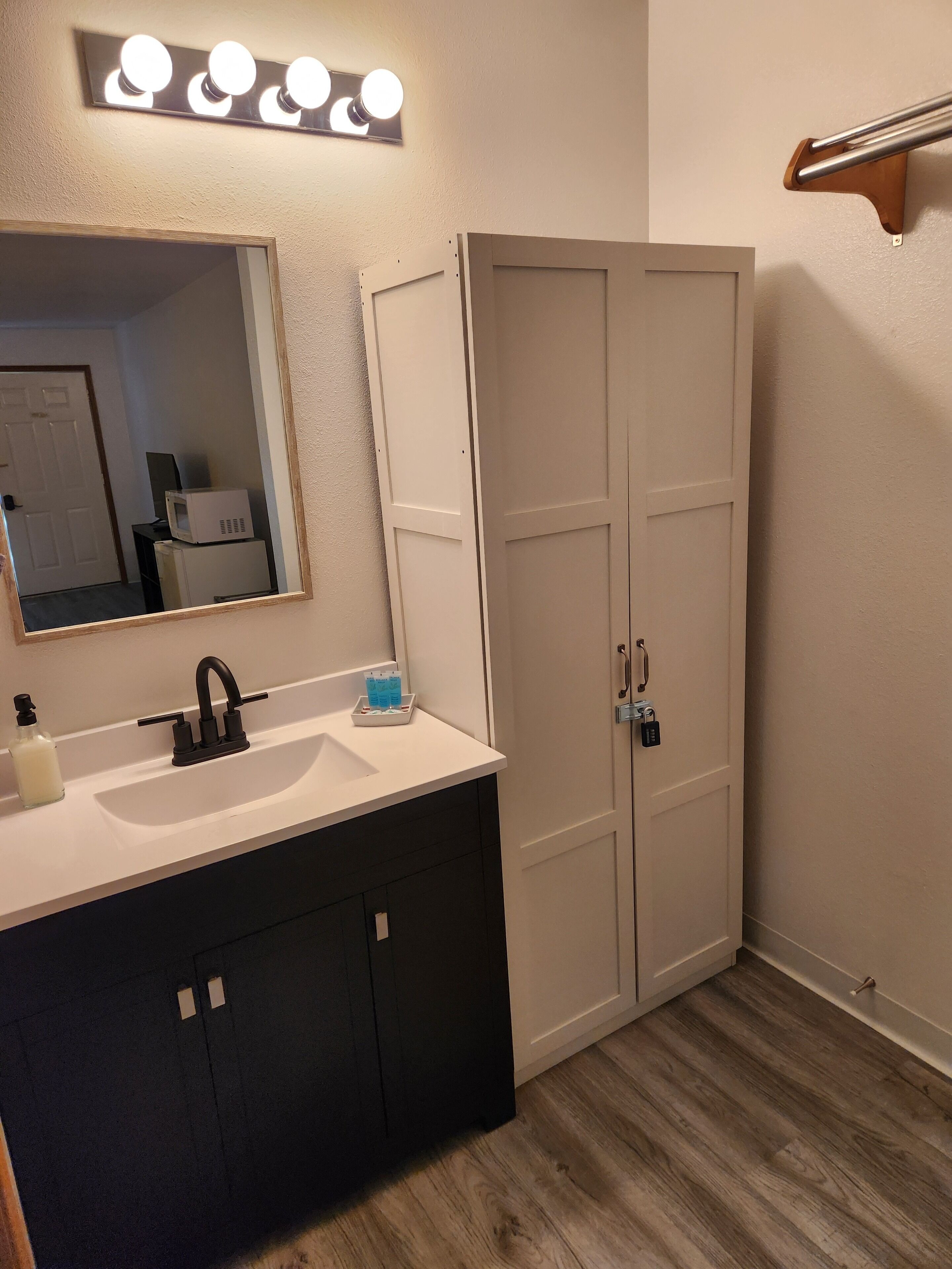 A bathroom with a white sink, dark cabinets, a mirror with lights, and a white door.