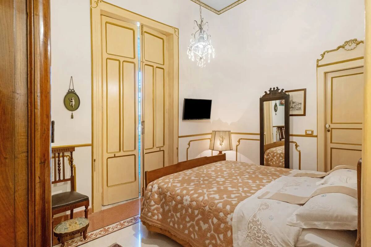 A bedroom with a large bed, a wooden chair, a small table, a television, and a chandelier.