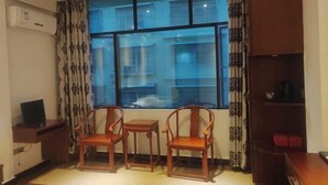 Een kamer met een groot raam, twee houten stoelen, een kleine tafel, een televisie op een standaard en een boekenplank.