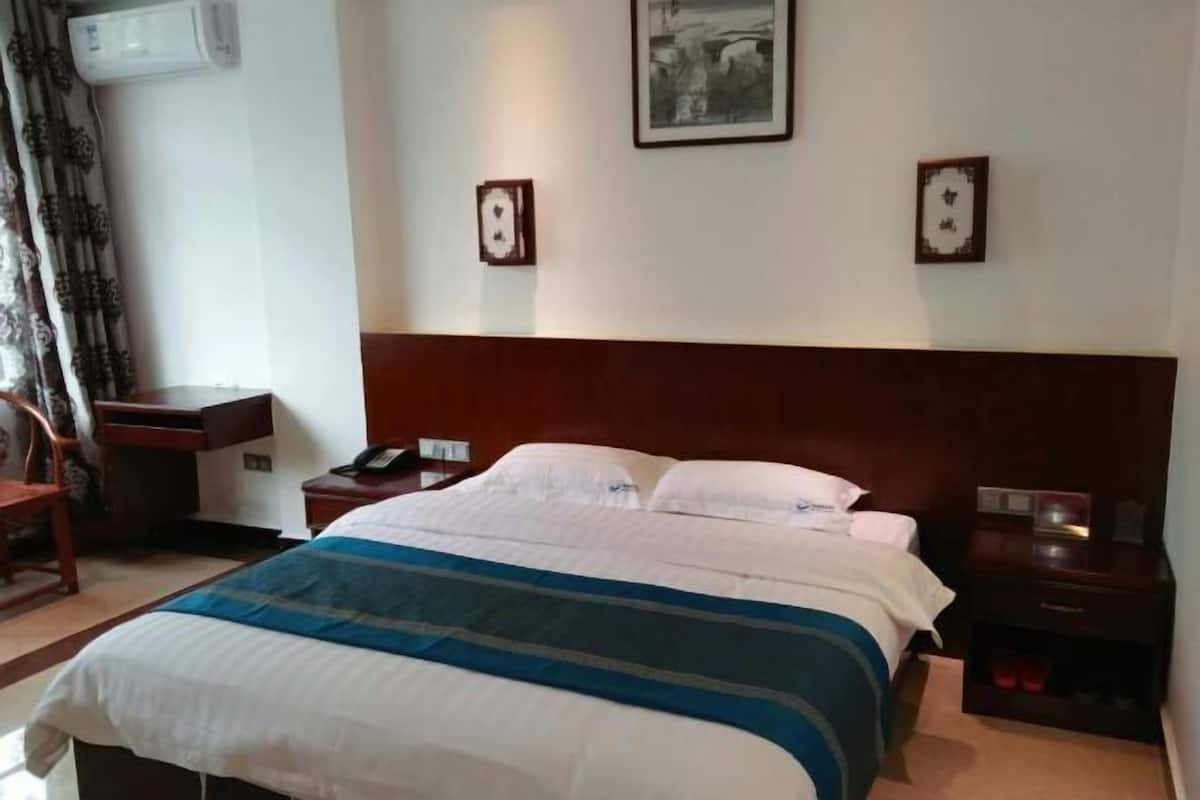 Hotellihuone, jossa on suuri sänky, puinen pääty, työpöytä tuolilla, yöpöytä ja seinälle asennettu ilmastointilaite.