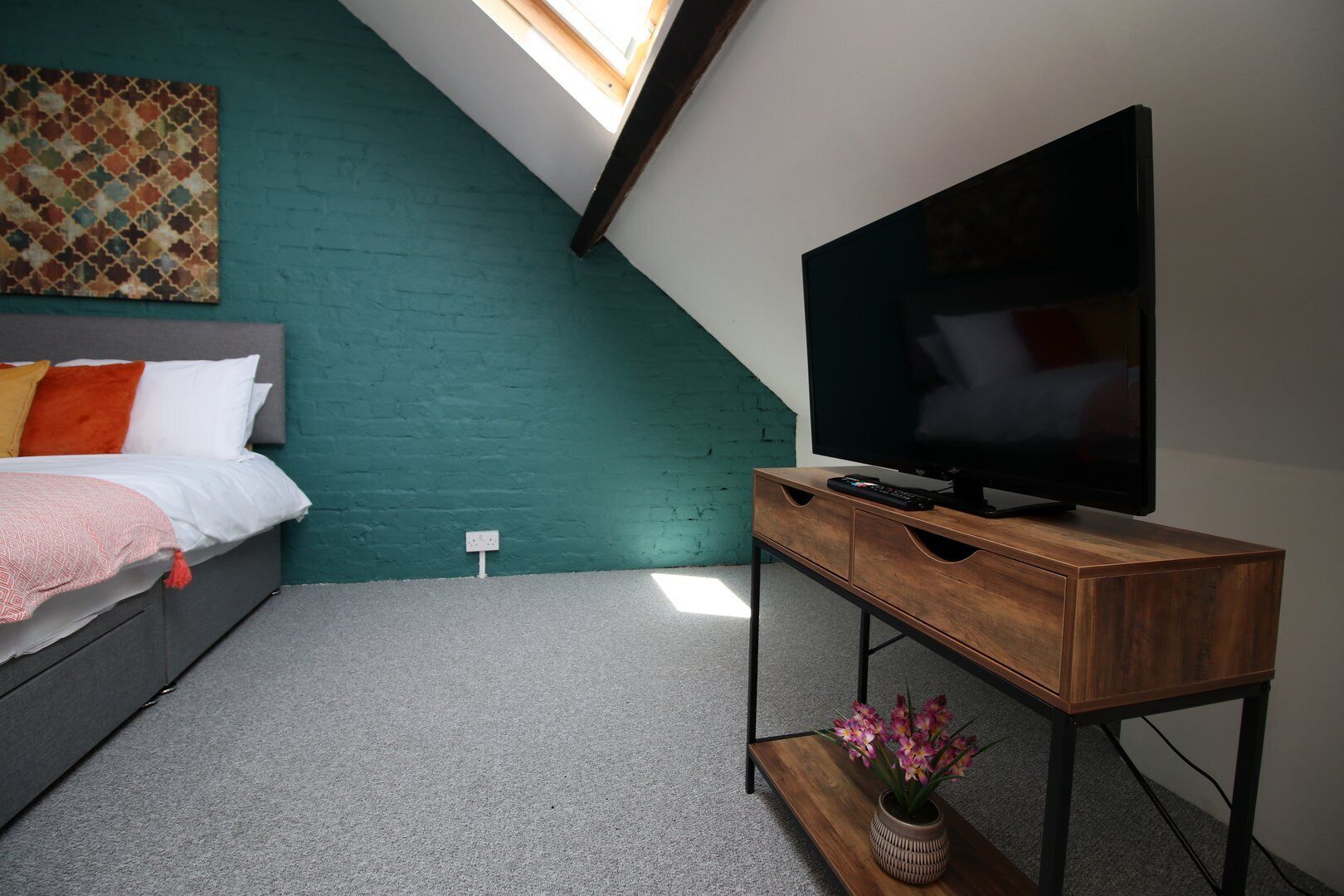 Une chambre à coucher avec un lit, un téléviseur posé sur un meuble en bois, une petite table avec des fleurs et un mur vert.