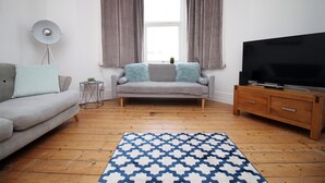 A living room with a grey sofa, a patterned rug, a TV on a wooden cabinet, and a window with curtains. - Cathedral House by Tŷ SA (Newport)