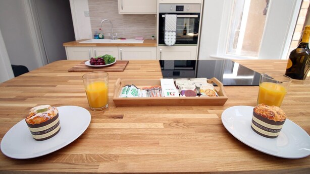 A breakfast setting with two plates of pastries, a tray of assorted snacks, and glasses of orange juice on a wooden table in a modern kitchen. - Cathedral House by Tŷ SA (Newport)