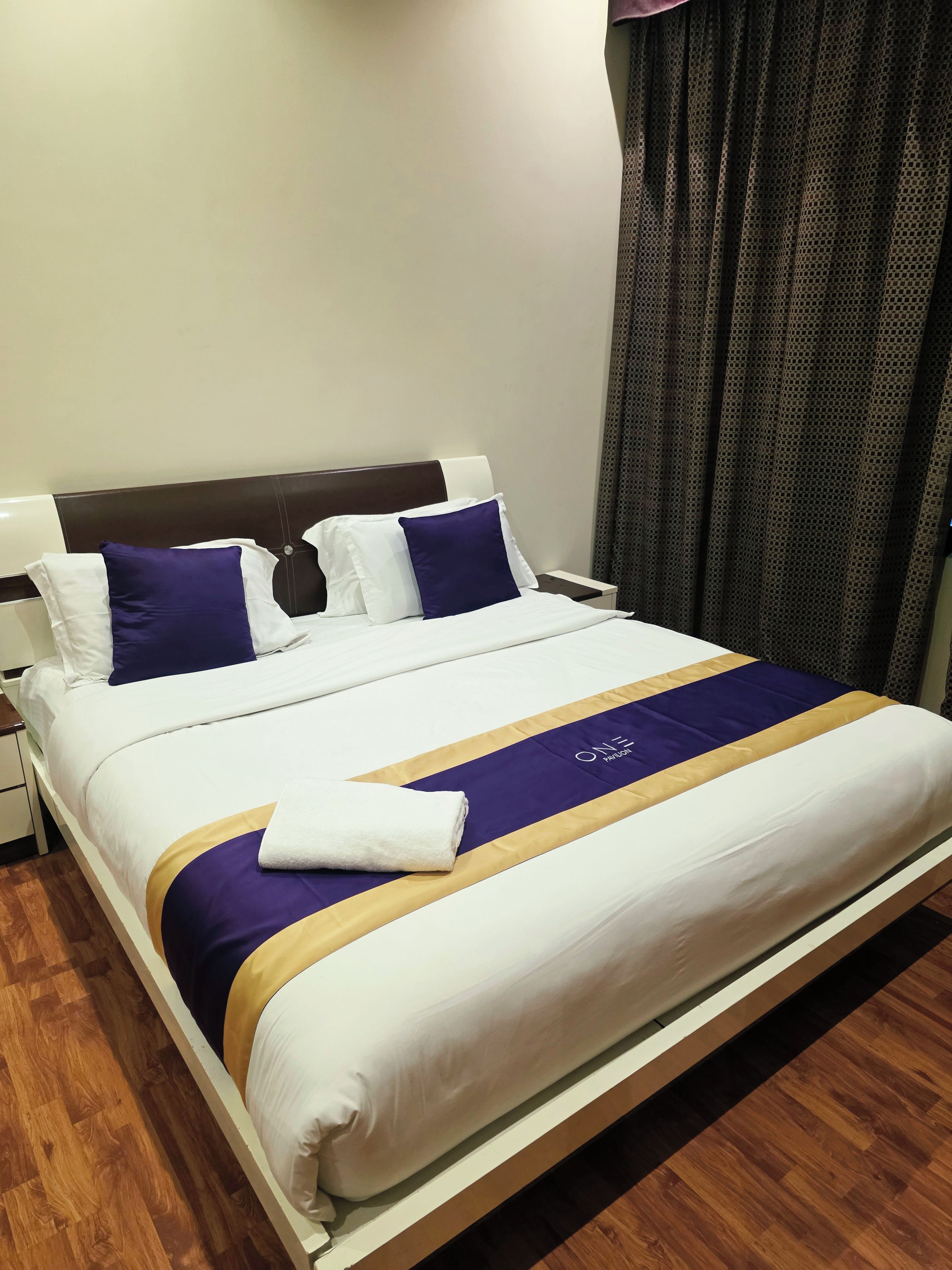 Een net opgemaakt bed met een wit-paarse spreidek en een opgevouwen witte handdoek, met een houten hoofdbord.