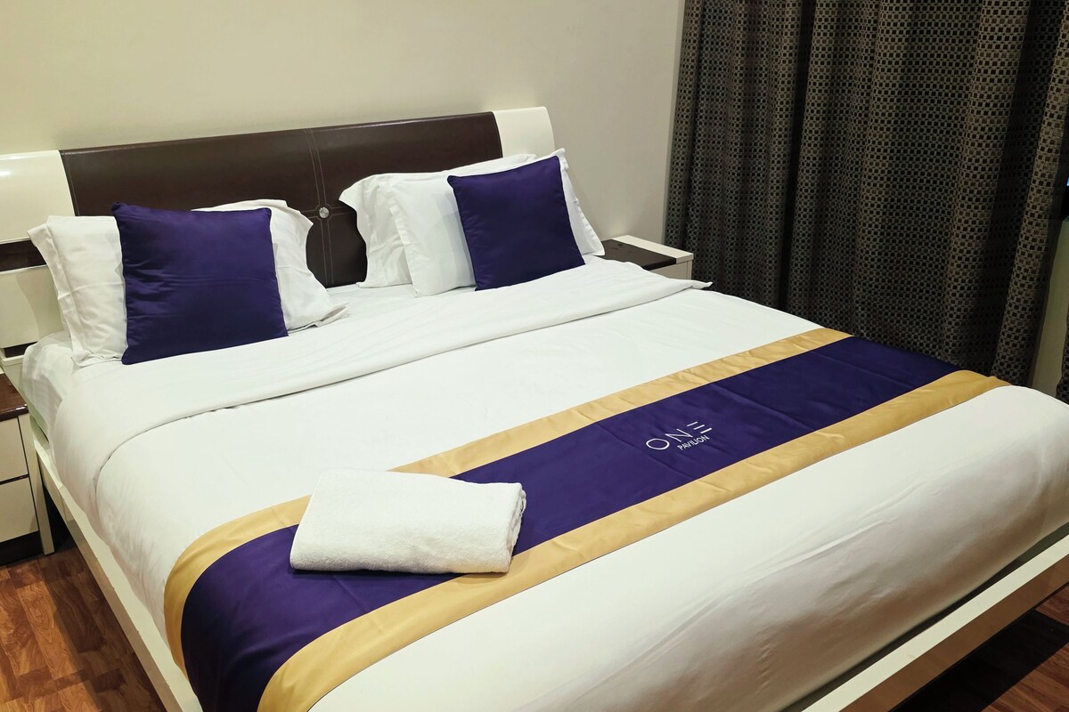 A neatly made bed with a white and purple bedspread, a folded white towel, and a wooden headboard.