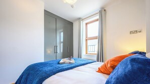 A bedroom with a bed, a blue blanket, an orange pillow, a window with curtains, and a wardrobe. - 2 bed with terrace - Llandaff Lofts by Tŷ SA (Cardiff)