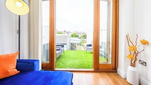 A room with a blue sofa, a floor lamp, a sliding glass door leading to a green lawn, and a white vase with yellow flowers. - 2 bed with terrace - Llandaff Lofts by Tŷ SA (Cardiff)