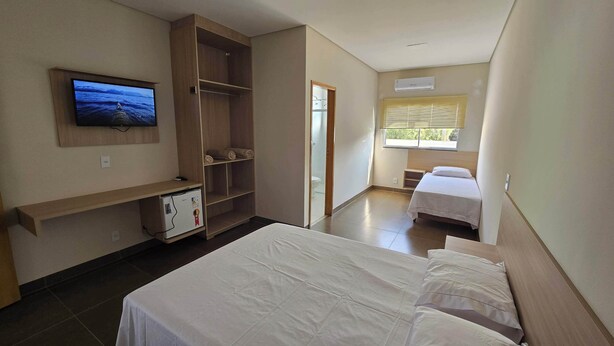 A hotel room with a bed, a TV mounted on the wall, a wardrobe, a small refrigerator, and a window with blinds. - Hotel Habitar (Serra do Salitre)