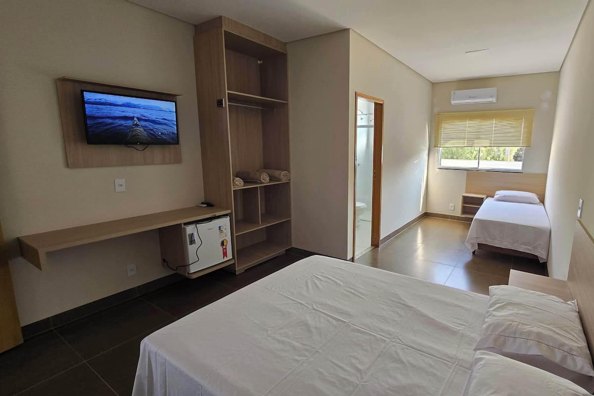 A hotel room with a bed, a TV mounted on the wall, a wardrobe, a small refrigerator, and a window with blinds.