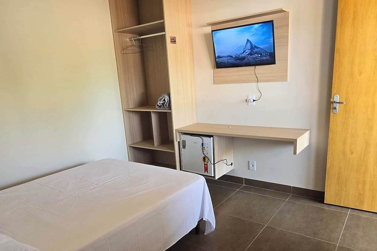 A hotel room with a bed, a TV mounted on the wall, a small refrigerator, and a wooden door.