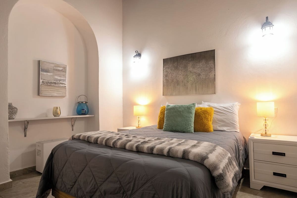 A bedroom with a bed, bedside lamps, a nightstand, a wall-mounted artwork, and a shelf with decorative items.