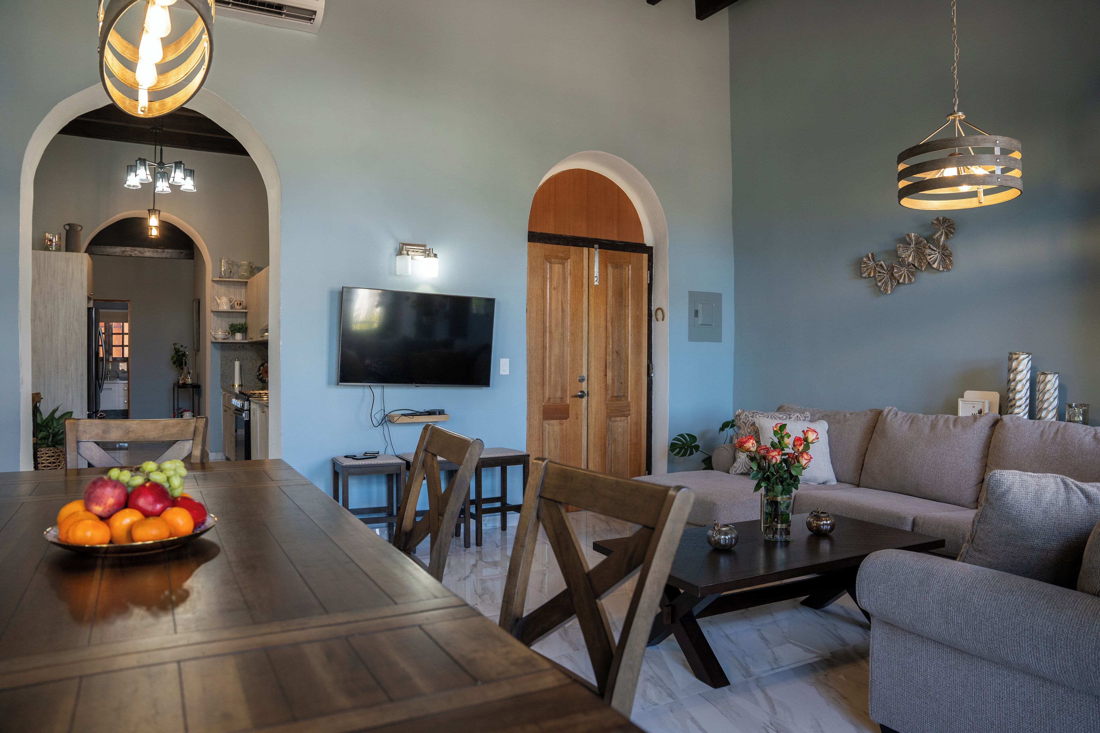 A dining area with a wooden table, chairs, and a fruit bowl. There is a television on the wall and a sofa in the background.