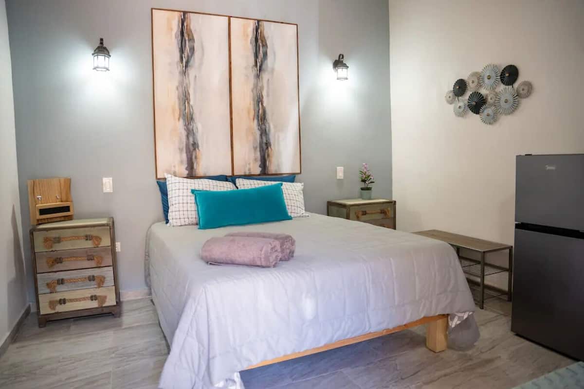 A bedroom with a bed, a dresser, a nightstand, a lamp, and a refrigerator.