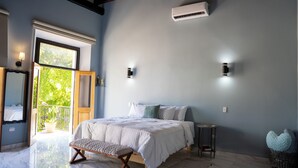 A bedroom with a bed, a bench, a small table, a wall-mounted air conditioner, and a view of greenery outside. - Hugo's Rental (San Juan)