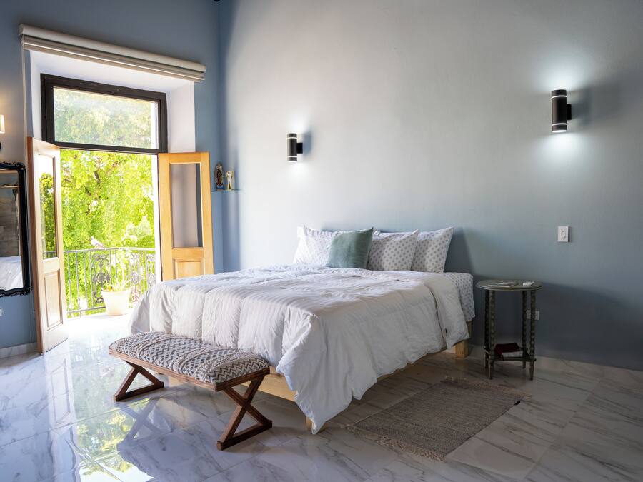 Quarto com cama grande, um banco, uma mesinha e vista para o exterior.