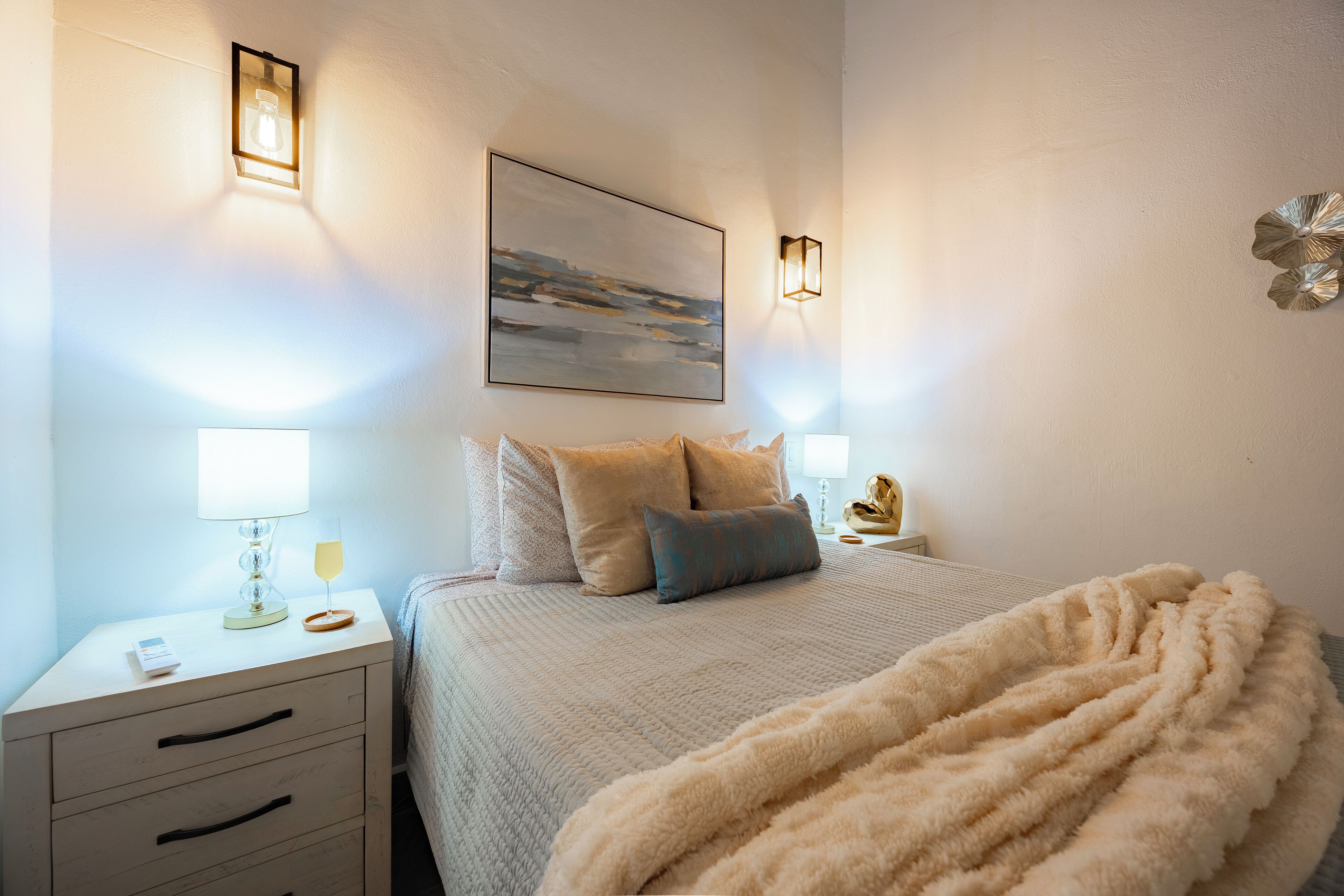 A neatly arranged bedroom with a bed, bedside tables, lamps, and a painting on the wall.