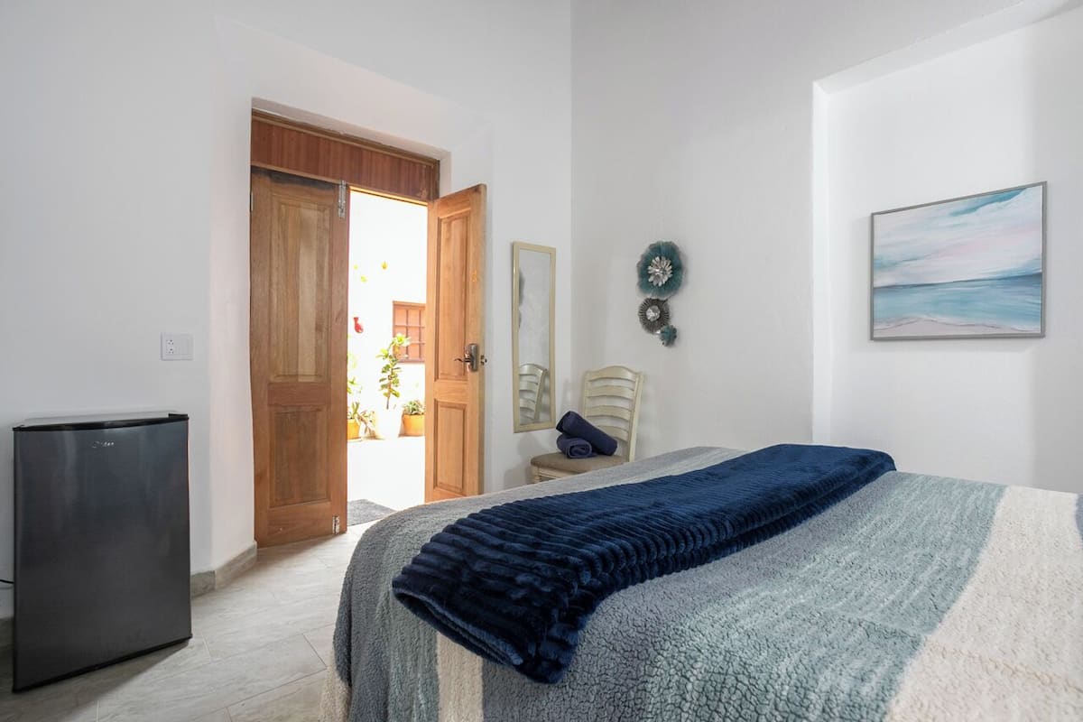 A bedroom with a bed, a small table, a chair, a door, and a refrigerator.
