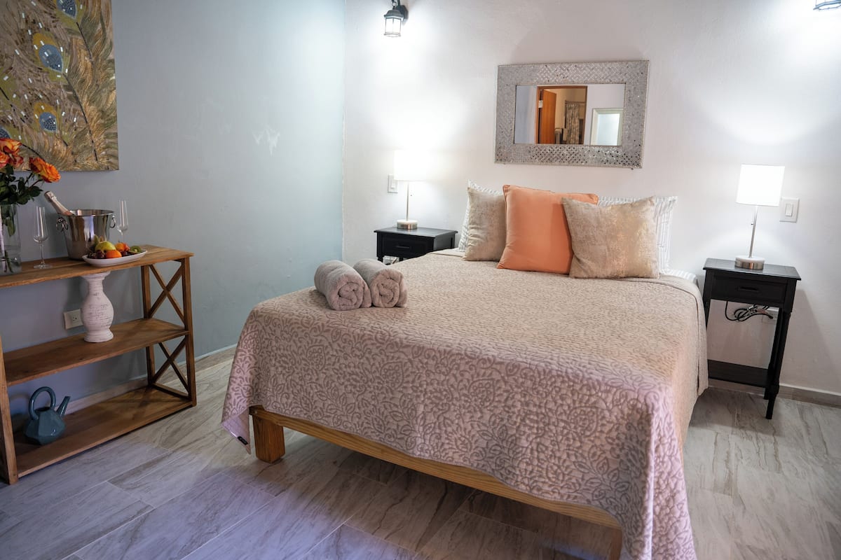 A neatly arranged bedroom with a bed, bedside tables, a nightstand, a wooden shelf, and a vase with flowers.