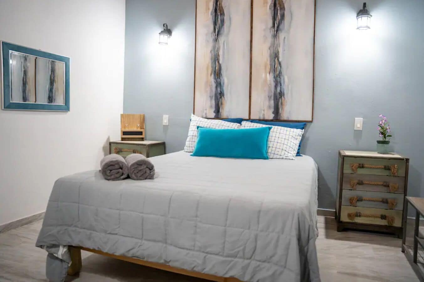 Una cama bien hecha con colcha blanca, dos toallas enrolladas y una almohada azul. Se observa un pequeño tocador de madera con una lámpara y un objeto decorativo encima.