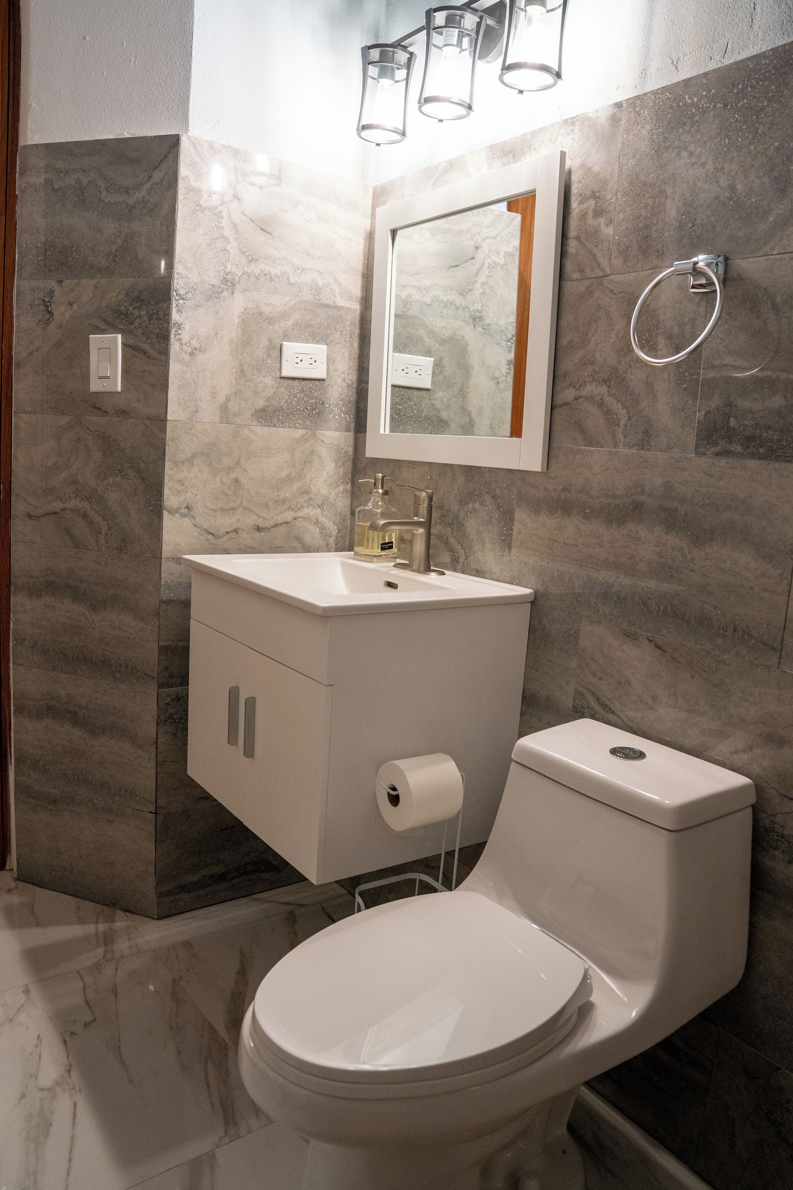 Un baño moderno con pared de mármol, un lavamanos blanco, un inodoro y un espejo con luces arriba.