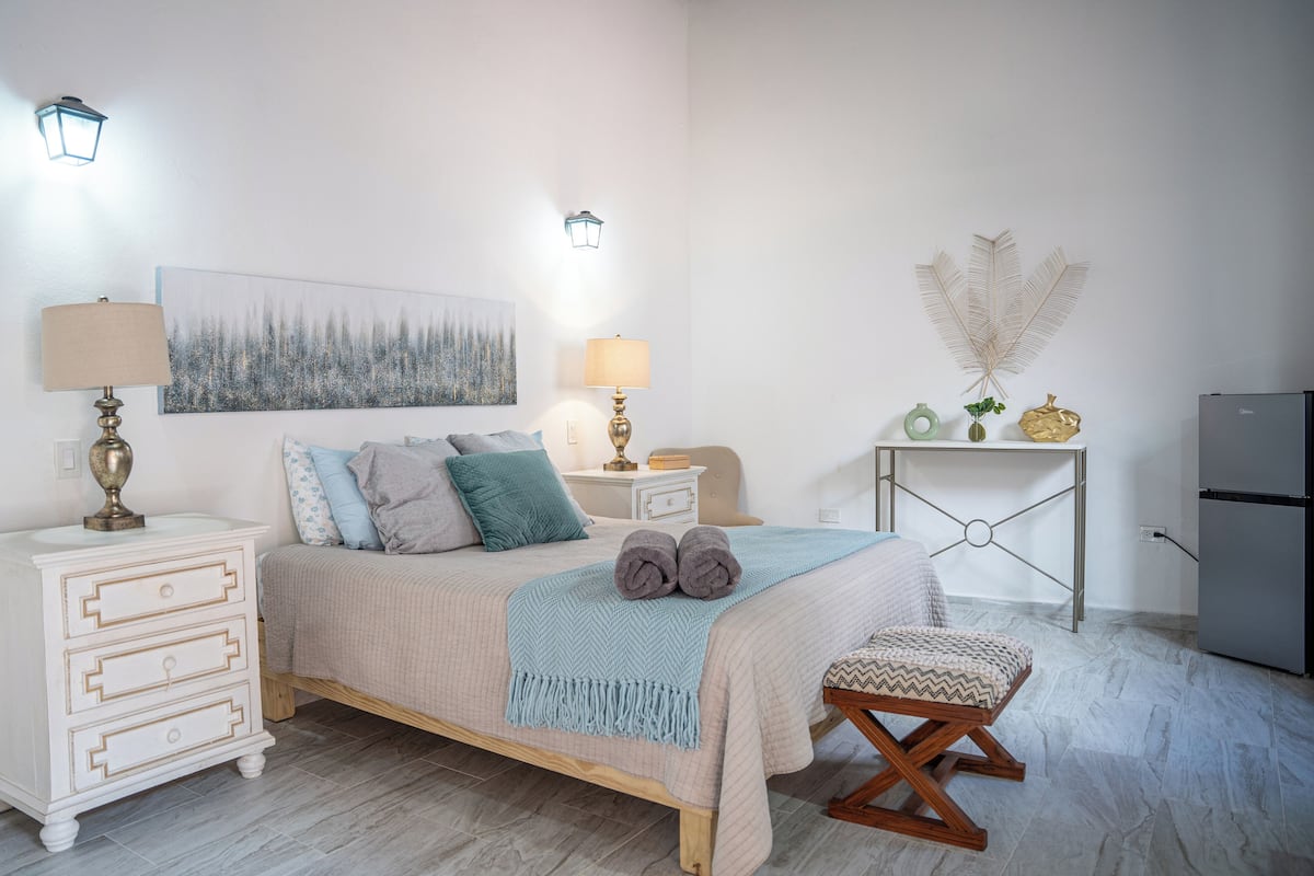 A bedroom with a bed, bedside tables, a lamp, a wall art piece, a small table with decorative items, and a refrigerator.