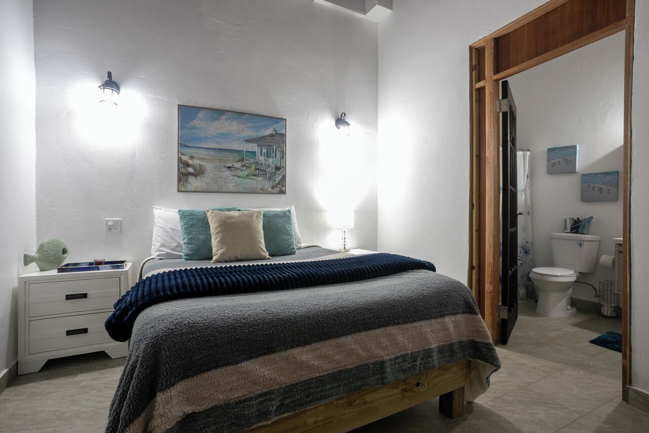 A bedroom with a bed, bedside tables, a painting, and a bathroom visible through an open door.