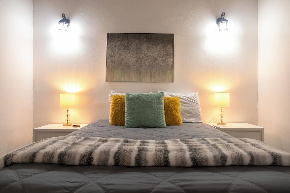 A neatly made bed with a quilted cover, flanked by two bedside lamps and a wall-mounted abstract painting.