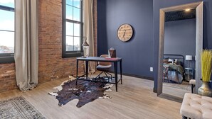 Habitación moderna con pared de ladrillo, un ventanal con cortinas, un reloj de pared redondo, una mesa con una silla, un tapete de cuero de vaca y un espejo que refleja una cama y una mesita de noche.