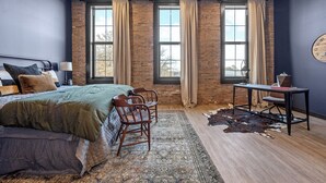 Un dormitorio con una cama grande, un escritorio y una silla. Hay tres ventanas con cortinas, una pared de ladrillo y un techo con diseño de cuadrícula.