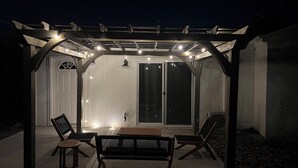 Un patio techado con un pérgola, dos bancos de madera y luces colgantes.