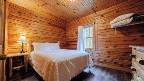 A cozy wooden cabin room with a bed, a nightstand with a lamp, a window with curtains, and a wooden dresser.