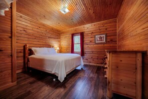 Quarto de cabana de madeira com uma cama, cômoda e janela com cortinas vermelhas.