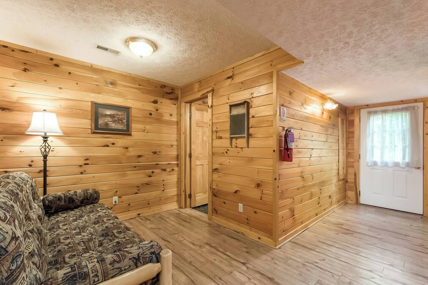 Una habitación con paneles de madera, un sofá, una lámpara, un cuadro enmarco, una puerta y un extintor.