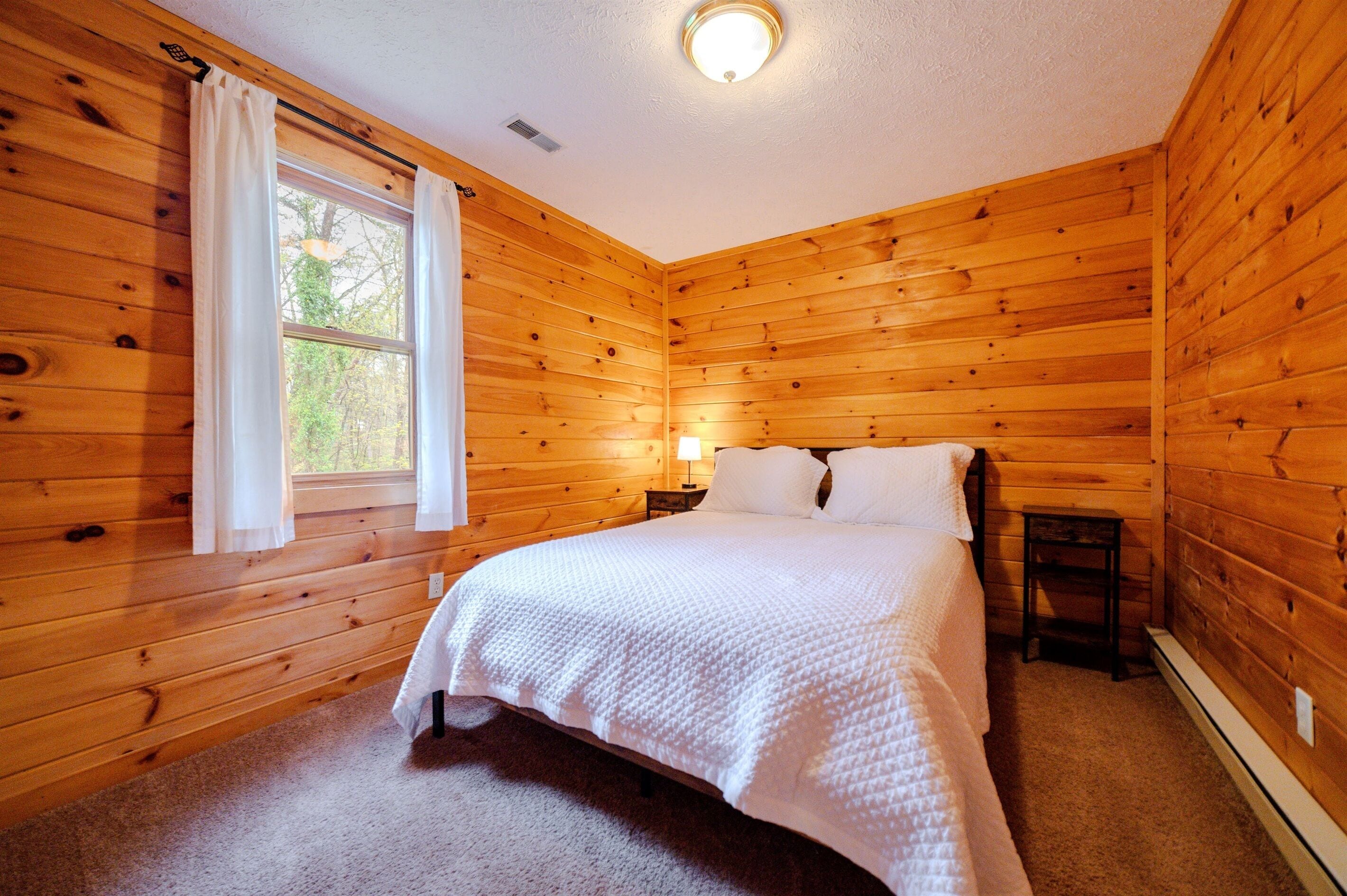 Un dormitorio con paneles de madera, una cama, una ventana con cortinas, una mesita de noche y una lámpara.