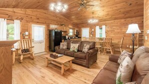 A cabin interior with wooden walls and floors, a black refrigerator, a brown sofa, a wooden coffee table, and a ceiling fan. - Family & Friends Getaway |Near Old Man's Cave (Logan)