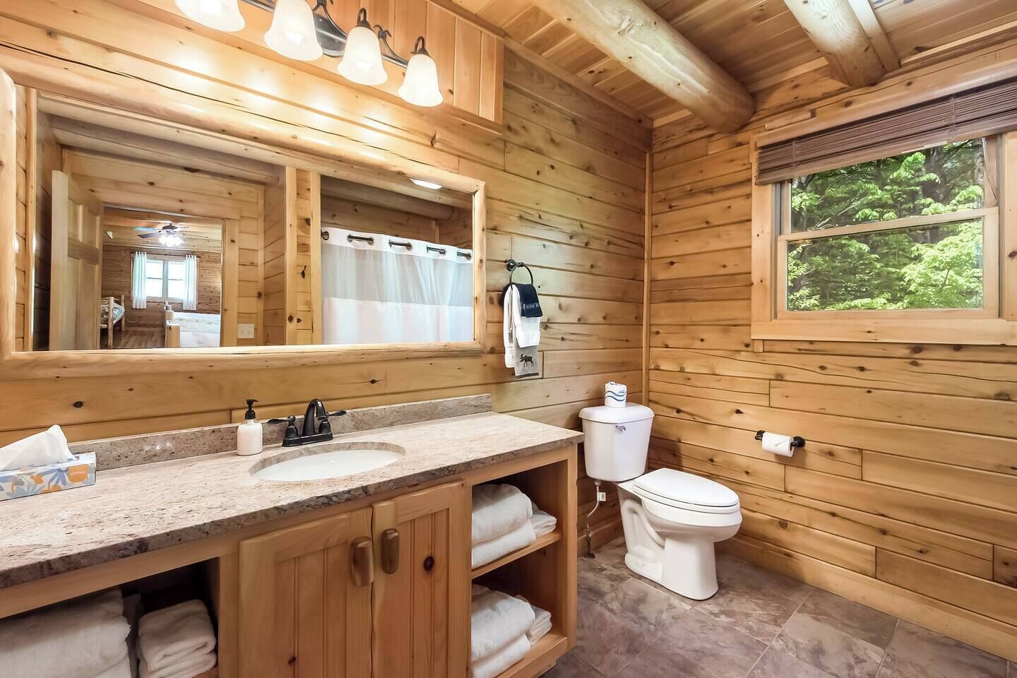 A wooden cabin bathroom with a large mirror, two sinks, a toilet, and a window with a view of trees.