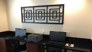 A hotel office with three desks, each with a laptop and a printer. There are two black office chairs and a decorative wall piece above the desks.