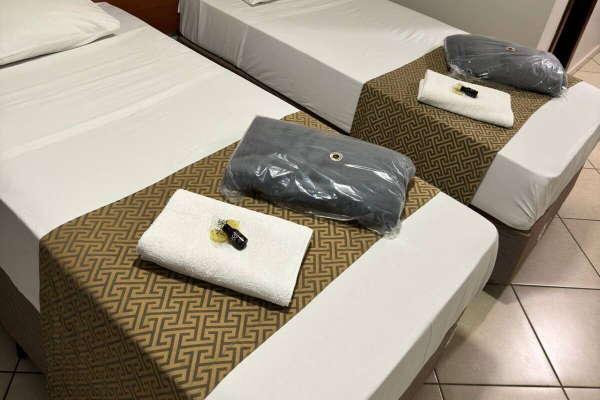 Quarto de hotel com duas camas, cada uma com roupa de cama branca e colcha estampada. Há um quadro na parede acima das camas.