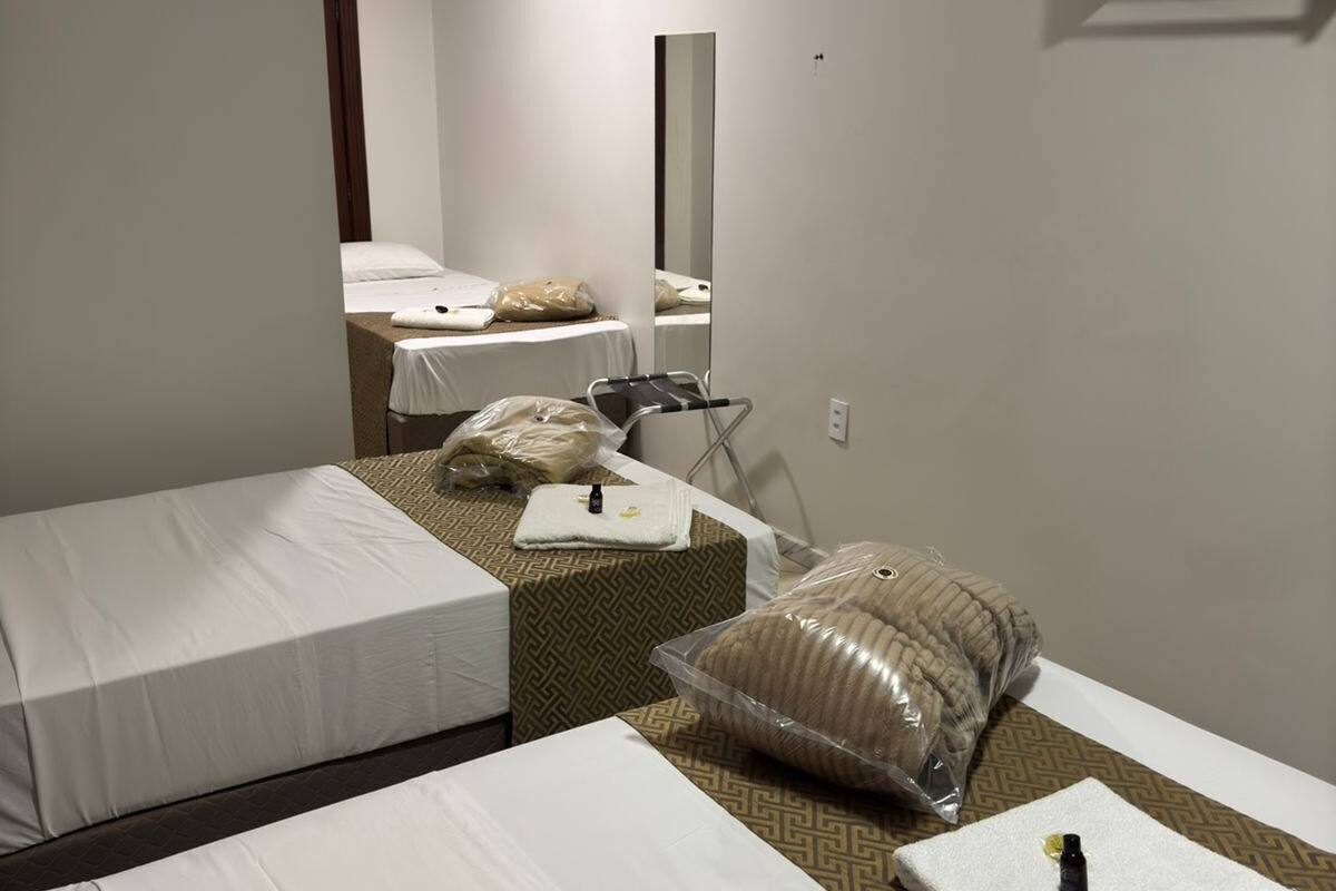 Quarto de hotel com duas camas de solteiro, uma mesinha, uma cadeira, um espelho e ar condicionado fixado na parede.