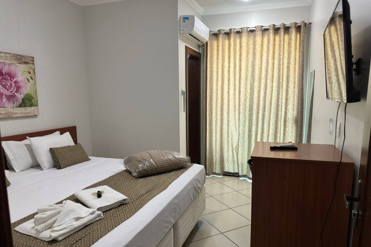 Quarto de hotel com cama, escrivaninha, televisão e chuveiro.