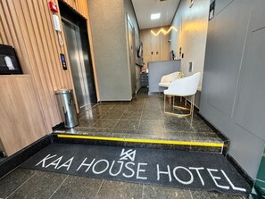 En hotellentrén med en svart dörrmatta där det står ”KAA HOUSE HOTEL” och en reception med modern design, inklusive en disk och sittplatser.